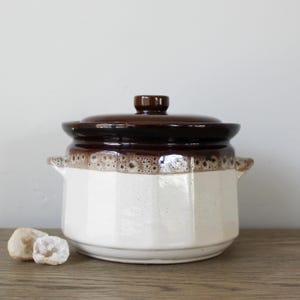 May include: A white ceramic crock pot with a brown lid. The crock pot has a brown band around the top and a white body with a textured pattern. The lid has a brown knob.