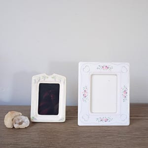 May include: Two decorative picture frames. One is rectangular with a floral design and the other is a smaller, arched frame with a lavender design. Both are white and sit on a wooden surface.