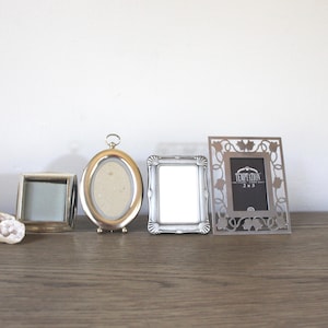 May include: Four decorative picture frames of varying shapes and sizes sit on a wooden surface. The frames are silver and gold-toned, with one featuring a floral design and the text "Temptation 2x3".