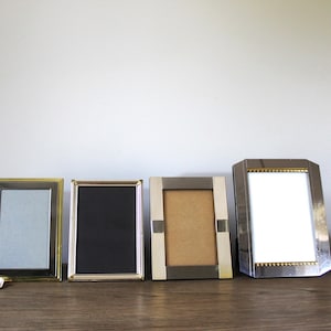 May include: Four decorative picture frames in a row, each with a different design. The frames are made of metal and have various colors, including gold, silver, and black. The frames are empty, with a light blue, black, and tan background.
