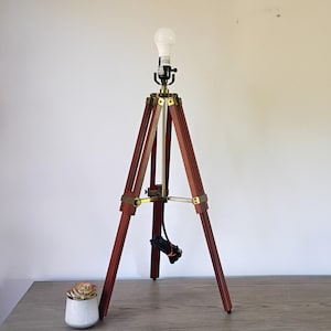 May include: A wooden tripod lamp with brass accents and a white lightbulb. The lamp has a dark brown finish and a classic design. A small succulent in a white pot sits nearby. The lamp's height is approximately 36 inches.