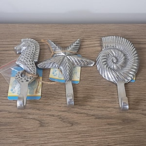 May include: Three silver-colored metal wall hooks. One is shaped like a seahorse, another like a starfish, and the third resembles a nautilus shell. Each hook has a sturdy, rectangular base for mounting.