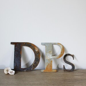 May include: Three metal letters, D, P, and S, are arranged in a row on a wooden surface. The letters are rusty and have a distressed look. The letters are all uppercase and are about 10 inches tall.