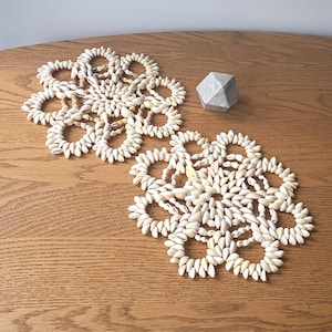 May include: Two decorative placemats made of small, cream-colored seashells arranged in a floral pattern. A geometric, gray stone object sits nearby on a wooden table. The placemats are circular with open centers.