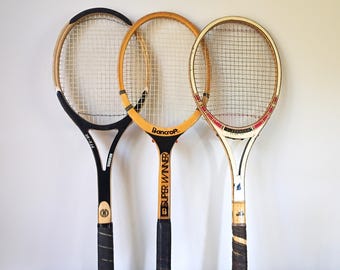 May include: Three vintage tennis rackets with wooden frames and string netting. One racket is black with a white and gold trim, another is yellow with "Bancroft Super Winner" text, and the third is white with red accents. The rackets have black grips.