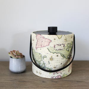 May include: A vintage-style ice bucket with a black lid and handles. The bucket is covered in a map print with a cream background and colorful accents. The map features the continents of North America, South America, Europe, Africa, and Asia.