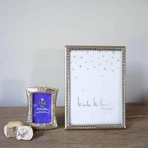 May include: A small silver-toned metal picture frame with a 2x3 inch insert and a larger rectangular silver-toned metal picture frame with a textured border and a 5x7 inch insert featuring a star pattern and the text Nicole Miller Home.
