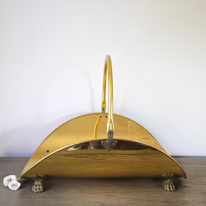 May include: A polished brass fireplace log holder with a curved body and a handle. The holder has decorative claw feet and is designed to hold firewood. The brass has a shiny, reflective surface.