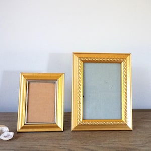 May include: Two rectangular picture frames with a metallic gold finish and a rope-style textured border. The smaller frame on the left contains a tan backing, while the larger frame on the right features a light blue backing within the display area.