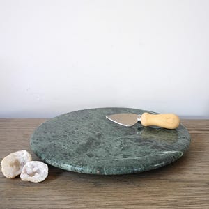 May include: A round, green marble cheese board with a stainless steel cheese knife and a light wood handle. Two small, white and beige geodes are in the foreground. The cheese board is on a wooden surface.