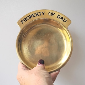 May include: A round, brass-colored dish with the words "PROPERTY OF DAD" in black lettering. The dish is held by a hand with a dark red nail polish. The dish has a slightly curved rim and a smooth, reflective surface.