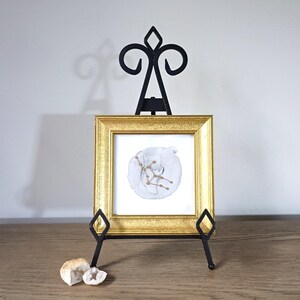 May include: A square gold-toned frame containing a white print with a grey circular watercolor wash and a gold constellation pattern. The frame is displayed on a black metal easel stand, with two small, rough-textured white geode stones nearby.