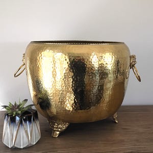 May include: A large, hammered gold metal bowl with three ornate feet and two ring handles. The bowl has a textured surface and a wide opening. A small, geometric planter with a succulent is next to the bowl.