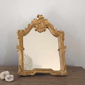 May include: A gold-colored, ornate mirror with a decorative frame. The mirror has a rectangular shape with a curved top and intricate carvings. The frame is detailed with floral and scrollwork designs, adding a touch of elegance.
