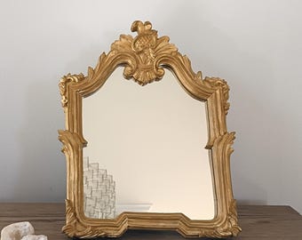 May include: A gold-colored, ornate mirror with a decorative frame. The mirror has a rectangular shape with a curved top and intricate carvings. The frame is detailed with floral and scrollwork designs, adding a touch of elegance.