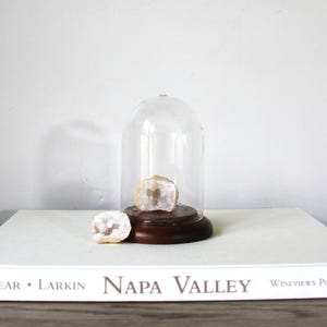 May include: A clear glass cloche with a wooden base houses a geode. Another geode sits beside the cloche on a white surface. The geode has a light brown exterior and a crystal interior. The word "Napa Valley" is visible on the book.