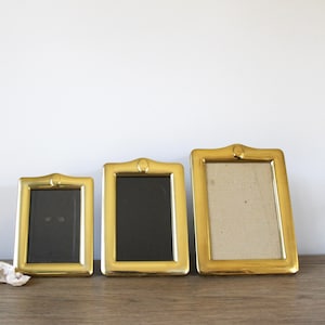 May include: Three gold-toned picture frames of varying sizes, each with a decorative design and a raised circular knob at the top.