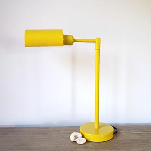 May include: A bright yellow desk lamp with a cylindrical shade and a round base. The lamp has a slender, adjustable arm and a matching yellow stem. The lamp is approximately 12 inches tall and is set on a wooden surface.