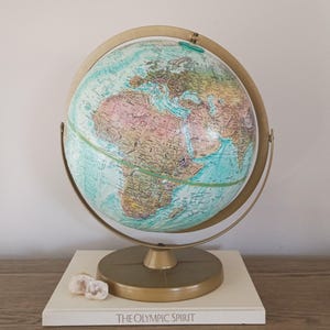 May include: A vintage globe with a gold-colored metal frame and base. The globe features a detailed map with various colors representing different countries and landforms. The base rests on a white book with the text "THE OLYMPIC SPIRIT".
