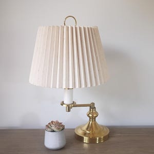 May include: A brass desk lamp with a pleated beige lampshade. The lamp has a curved arm and a round base. A small succulent in a white pot sits next to the lamp on a wooden surface.