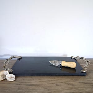 May include: A rectangular black stone serving board featuring two metallic handles with an organic, textured finish. A small cheese knife with a stainless steel blade and a light-colored wooden handle rests on the surface of the board.