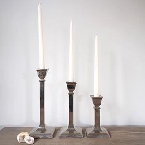 May include: Three silver-toned metal candle holders of varying heights, each holding a tall, white candle. The candle holders have a square base and a rectangular stem. The candles are unlit, and the background is a plain white wall.