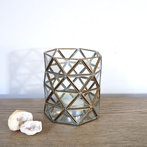 May include: A cylindrical candle holder constructed from clear glass panels framed in a metallic gold-toned metal with a geometric triangular lattice pattern. A small white tea light candle sits inside the base, and two small geode rocks are nearby.