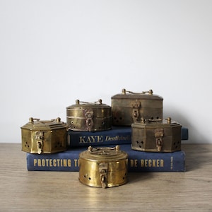 May include: Five antique brass boxes with intricate designs and latches. The boxes are stacked on top of two blue books with the text "KAYE Death in the" and "PROTECTING TI DE BECKER" visible.