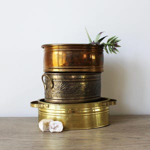 May include: Three brass planters, one with a copper top, stacked on top of each other. The top planter has a small green plant inside.