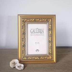 May include: A rectangular gold photo frame with an ornate, textured border and a smooth inner edge. The insert displays the text GALLERIA, 4 x 6 inches, and 10 cm x 15 cm. Two small, light-colored, crystalline geode rocks sit at the base of the frame.