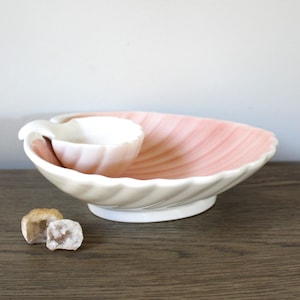 May include: A vintage ceramic serving dish and cup set. The dish is shell-shaped with a pink interior and white exterior. The cup is white and sits inside the dish. Two small, rough-cut stones are in front of the dish. The set is on a wooden surface.