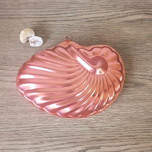 May include: A copper-colored, nautilus-shaped mold with a handle and the text "6 cups". The mold has a spiral design and is on a wooden surface. Two small, white, geode-like stones are next to the mold.