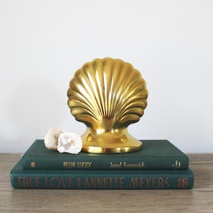 May include: A gold-colored seashell bookend with a smooth, shiny finish. The bookend is sitting on top of two green books. The top book has the title "Plum Lucky" and the author "Janet Evanovich". The bottom book has the title "Free Love" and the author "Jannette Meyers".