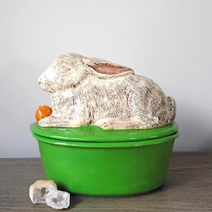 May include: A ceramic bunny figurine with a light brown and cream fur design, perched atop a bright green bowl. The bunny holds a small orange carrot. Two small, white, geode-like stones sit in front of the bowl.