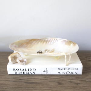 May include: A pearlescent, oval-shaped shell dish with small, gold-colored feet, holding a ring and a pearl necklace. The dish rests on a white book with the title "Rosalind Wiseman" and "Masterminds & Wingmen". Two geodes are on the left.