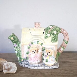 May include: A whimsical ceramic teapot shaped like a cottage, featuring two teddy bears in front of the door. The teapot has a light yellow roof, a chimney, and a tree-shaped handle. The spout and handle are adorned with green leaves and flowers.