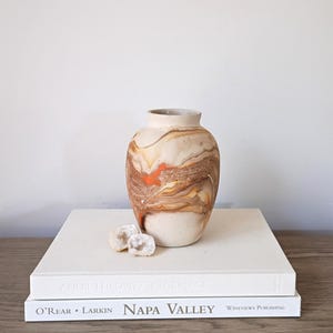 May include: A decorative ceramic vase with a marbled design in shades of beige, brown, and orange. The vase is displayed on a stack of white books, with two small, light-colored stones in front.