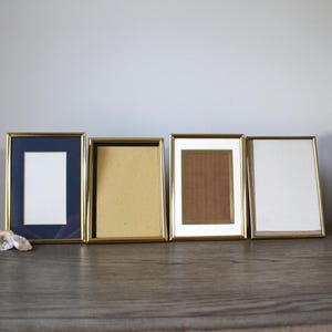 May include: Four rectangular picture frames with gold-colored metal edges. The frames have different colored matting, including navy blue, tan, brown, and white. The frames are displayed on a wooden surface.