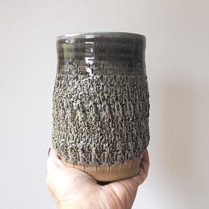 May include: A handmade ceramic vase with a textured surface. The vase has a dark brown top that fades into a textured, gray-brown body. The base is a lighter tan color. The vase is held in a hand against a white background.