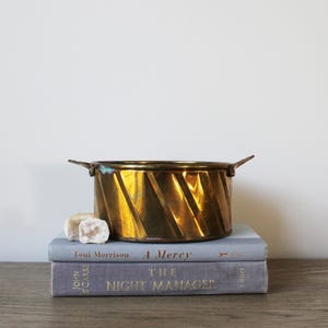 May include: A gold metal planter with a twisted design and two handles. The planter is sitting on top of two books, one titled "A Mercy" by Toni Morrison and the other titled "The Night Manager" by John le Carre.