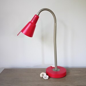 May include: A desk lamp featuring a vibrant red conical shade and a matching circular red base. The lamp has a flexible, metallic silver-toned gooseneck arm that allows for adjustable positioning of the light source, providing a functional design.