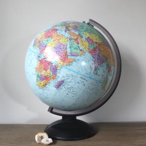 May include: A colorful globe with a light blue ocean and multi-colored continents. The globe is mounted on a black base with a dark gray meridian ring. A small, white, geode is in front of the globe.