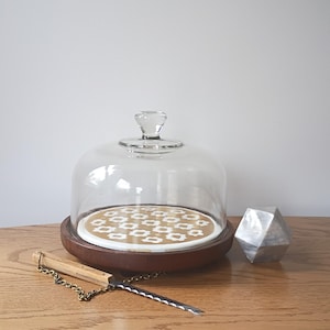 May include: A clear glass cake dome with a glass handle sits on a brown wooden base. A decorative silver geometric object and a cake knife with a wooden handle and chain are also present.