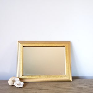 May include: A rectangular gold-framed mirror with a ridged border. The mirror is resting on a wooden surface, with two small, white, geode stones to the left. The mirror reflects the light, creating a warm, inviting aesthetic.