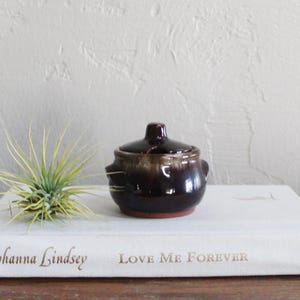 May include: A small brown and white ceramic pot with a lid. The pot has a rustic look and is sitting on a white book with the words "Love Me Forever" printed on it.
