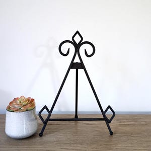 May include: Black metal display easel with decorative scrollwork and diamond accents. The three-legged easel is designed to hold a small framed picture or decorative plate. A small succulent plant in a white ceramic pot sits to the left of the easel.