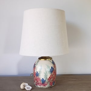 May include: A ceramic table lamp with a cream-colored lampshade. The base is white with a floral design in red, blue, and green. The lamp has a gold-colored band at the top of the base. Two small, light-colored stones are in front of the lamp.