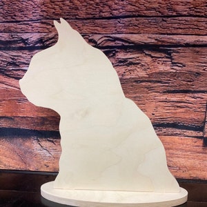 May include: A wooden cutout of a cat in profile, standing on a round base. The cutout is unpainted and made of light-colored wood.