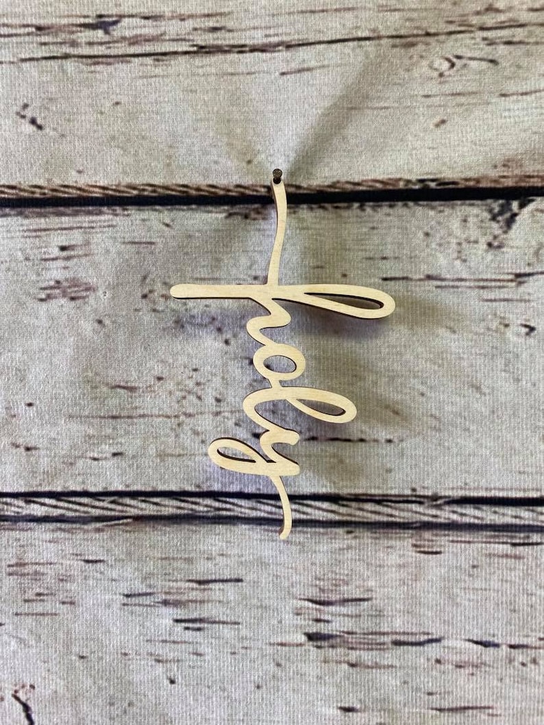 K&ouml;nnte beinhalten: Ein Holzausschnitt des Wortes "holy" in einer kursivschriftlichen Schriftart. Der Ausschnitt ist hellbraun und h&auml;ngt an einem Nagel auf einem hellbraunen Holzhintergrund.