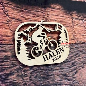 May include: A wooden ornament featuring a silhouette of a person riding a mountain bike through a mountain landscape. The ornament is personalized with the name "HALEN" and the year "2020".